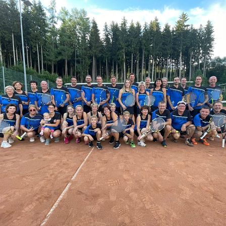 Eine Gruppe von Menschen in blauen Uniformen posiert für ein Foto auf einem Tennisplatz mit Bäumen im Hintergrund.