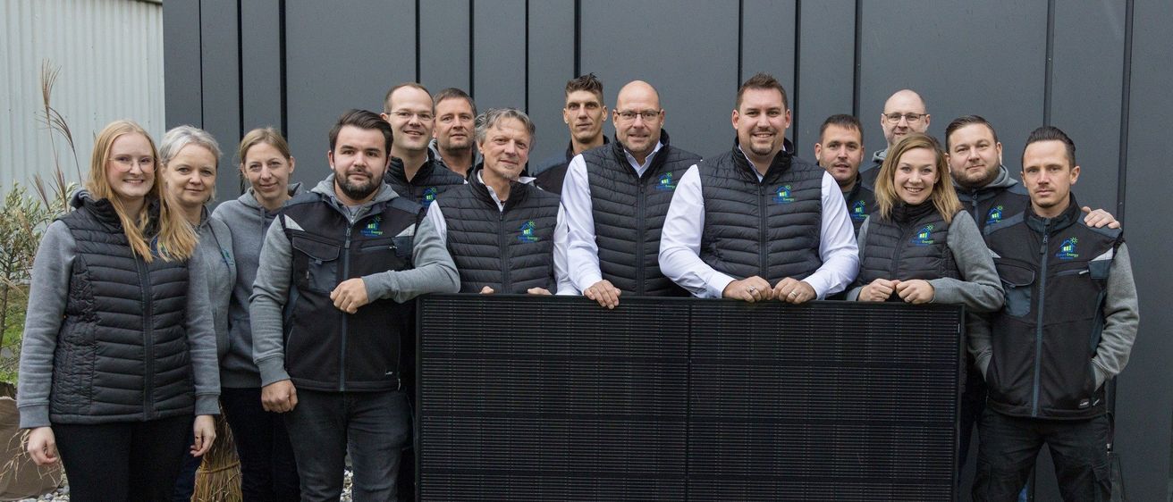 Eine Gruppe von Menschen in Jacken posiert für ein Foto vor einem schwarzen Solarpanel. Sie lächeln und scheinen Teil eines Teams zu sein.