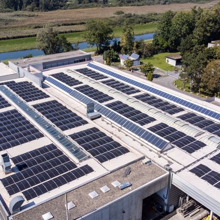 Eine Draufsicht auf ein großes Industriegebäude mit einem mit Solarpanelen bedeckten Dach, in der Nähe eines Flusses und umgeben von Bäumen.