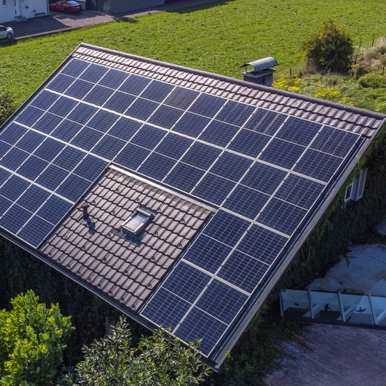 Eine Luftaufnahme eines Hauses mit einem mit Solarmodulen bedeckten Dach. Auf dem Dach befindet sich ein Schornstein. Das Haus ist von einem grünen Feld umgeben.