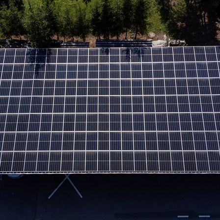 Eine Draufsicht auf eine Solarpanel-Anlage mit vielen ordentlich angeordneten Solarmodulen. Die Module sind dunkel und glänzend und reflektieren Licht. Sie sind auf einer weißen Struktur auf einer gepflasterten Oberfläche platziert.