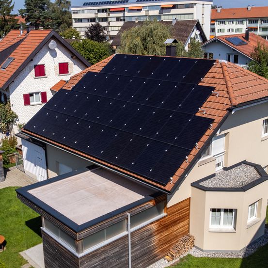 Ein Luftbild eines modernen Hauses mit Solarpanelen auf dem Dach, umgeben von anderen Häusern und Bäumen. Das Haus verfügt über ein Ziegeldach, weiße Wände und Holzakzente.