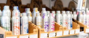 Ein Regal gefüllt mit verschiedenen silbernen Flaschen, jede mit dem Markennamen 'SAL' beschriftet. Die Flaschen sind ordentlich in Kartonboxen mit Etiketten organisiert. Der Hintergrund zeigt eine Wand mit einer hölzernen Struktur.