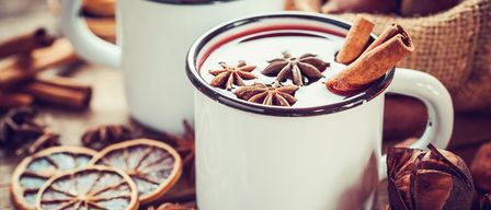 Zwei weiße Tassen mit roter Flüssigkeit, Sternanis und Zimtstangen. Im Hintergrund befinden sich getrocknete Orangenscheiben.
