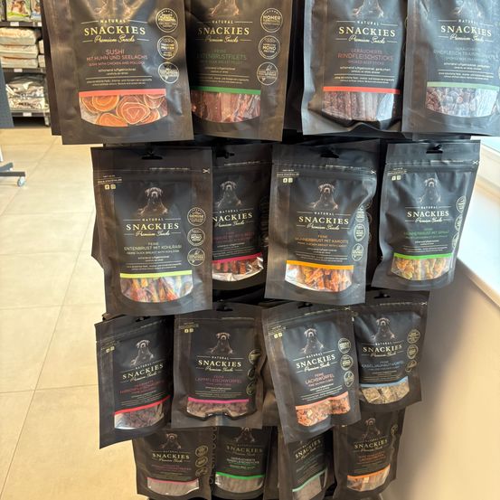 Eine Ausstellung verschiedener 'Natural Snackies' Hundesnacks, einschließlich Hühnchen- und Rindfleischgeschmack, auf einem Regal in einem Geschäft geordnet.