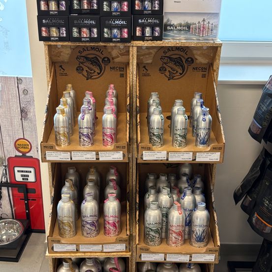 Eine Anzeige von Salmonflaschen in verschiedenen Farben und Designs. Die Flaschen sind in zwei Holzkisten mit Salmon-Marke angeordnet. Über den Kisten befinden sich Schachteln mit Salmon-Dosen.