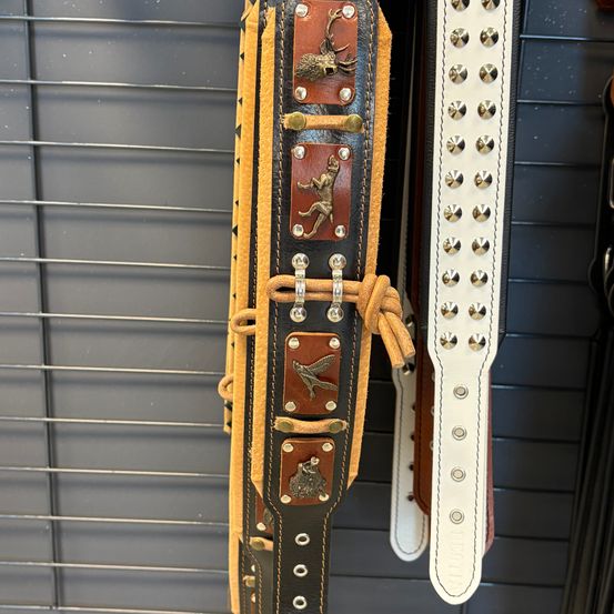 Ein brauner Ledergürtel mit Hirsch- und Froschdesigns hängt an einem Metallgitter, neben einem weißen Ledergürtel mit Nieten.