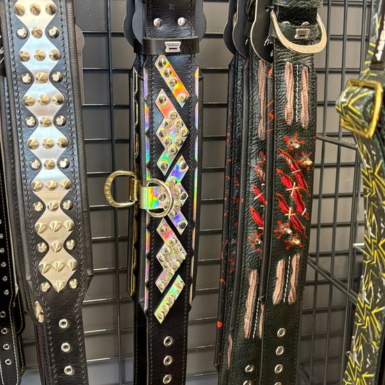 Eine Ausstellung von schwarzen Ledergürteln mit verschiedenen Designs, einschließlich Nieten, geometrischen Mustern und roter und weißer Stickerei, auf einem Metallständer.