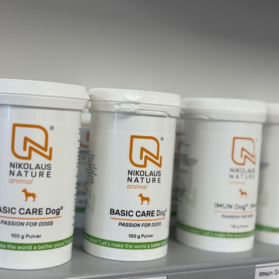 Mehrere weiße Behälter mit Nikolaus Nature Animal Basic Care Dog Pulver sind auf einem Regal aufgereiht, jeder mit einem grünen Streifen am Boden.