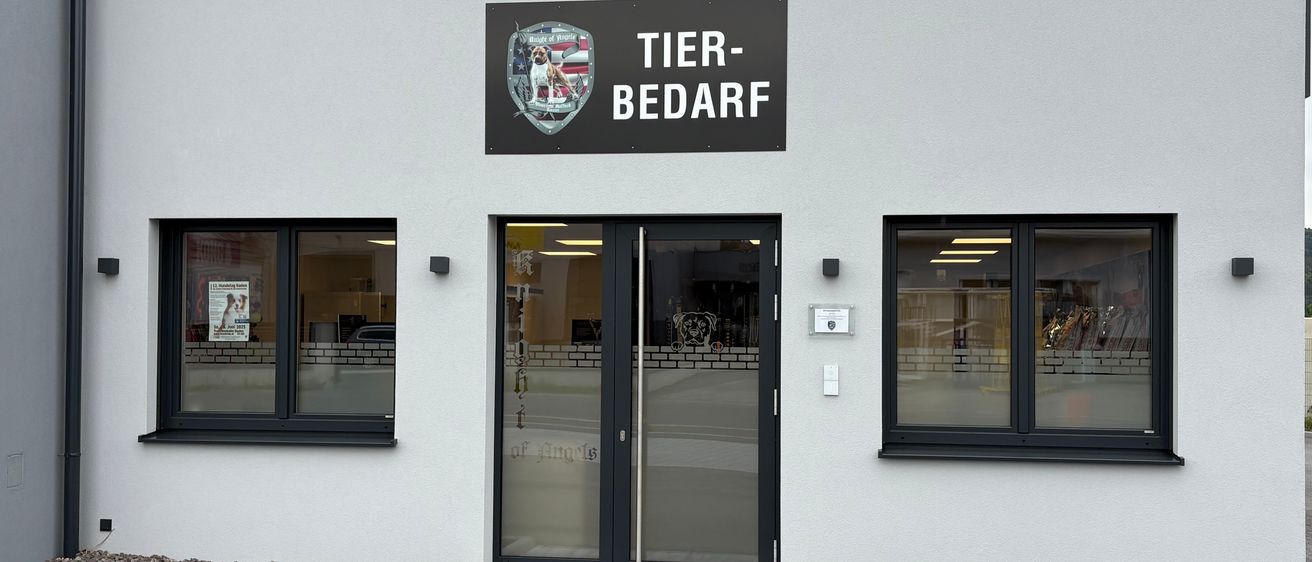 Das Äußere eines weißen Gebäudes zeigt ein großes Schild mit der Aufschrift 'TIER-BEDARF'. Unter dem Schild befinden sich Doppelglastüren und drei Fenster.