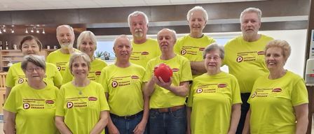 Eine Gruppe älterer Menschen in gelben T-Shirts mit Text und Logos posiert für ein Foto auf einer Treppe. Der Mann in der Mitte hält einen roten Bowlingball.