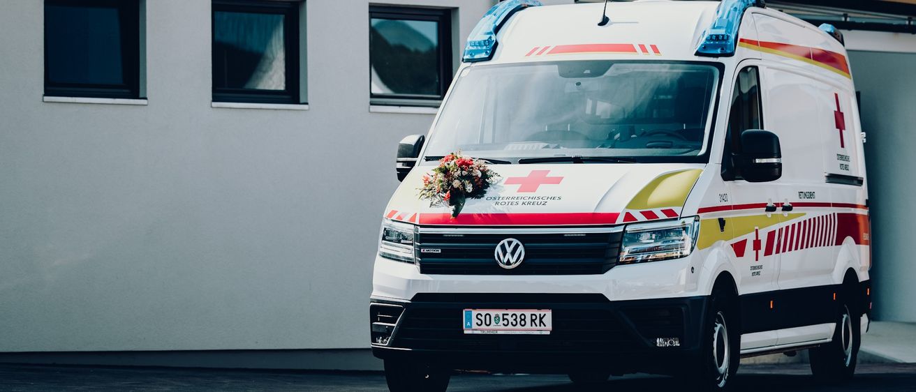Ein Krankenwagen mit einem Blumenstrauß auf der Front ist vor einem Gebäude geparkt. Der Krankenwagen hat ein rotes Kreuz auf der Front und ein Nummernschild SO 538 RK.