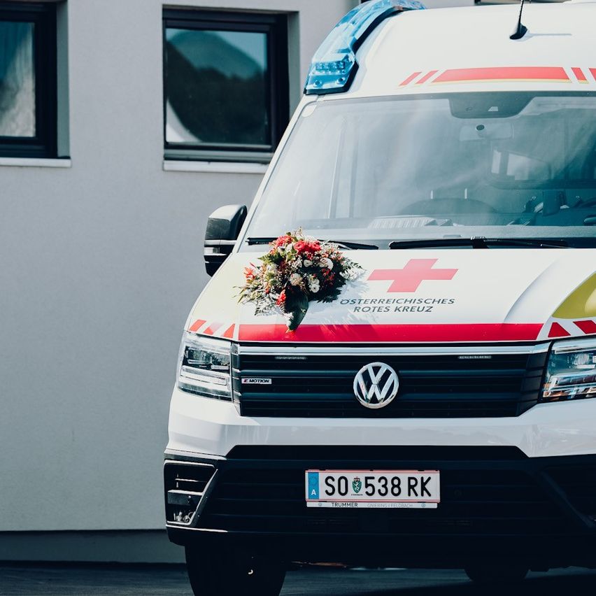 Ein Krankenwagen mit einem Blumenstrauß auf der Front ist vor einem Gebäude geparkt. Der Krankenwagen hat ein rotes Kreuz auf der Front und ein Nummernschild SO 538 RK.