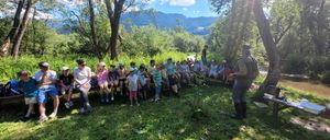 Eine Gruppe von Kindern und Erwachsenen sitzt auf einer Bank im Wald, mit Bergen im Hintergrund.