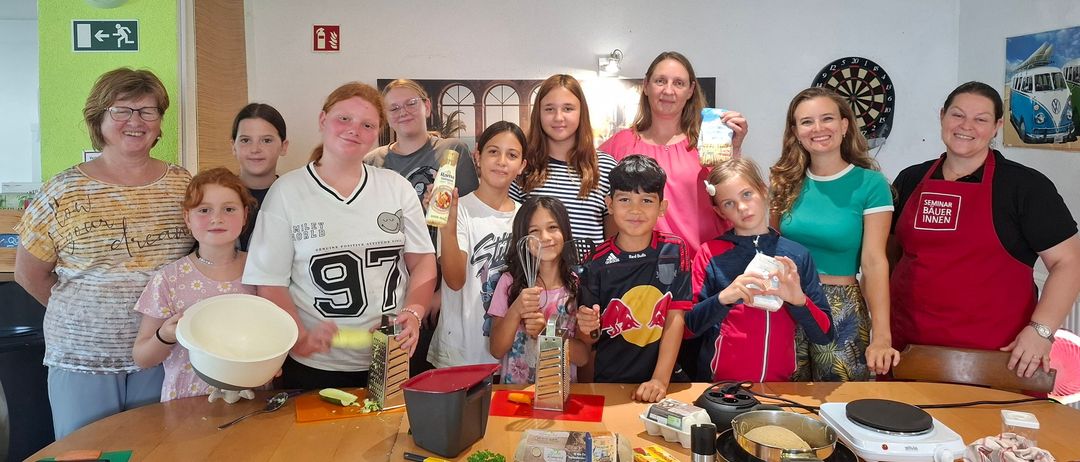 Eine Gruppe von Kindern und Erwachsenen versammelt sich um einen Tisch in einer Küche, hält verschiedene Küchenutensilien und Zutaten in den Händen. Sie lächeln und posieren für ein Foto.