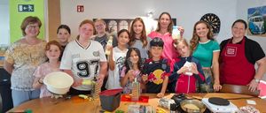 Eine Gruppe von Kindern und Erwachsenen versammelt sich um einen Tisch in einer Küche, hält verschiedene Küchenutensilien und Zutaten in den Händen. Sie lächeln und posieren für ein Foto.