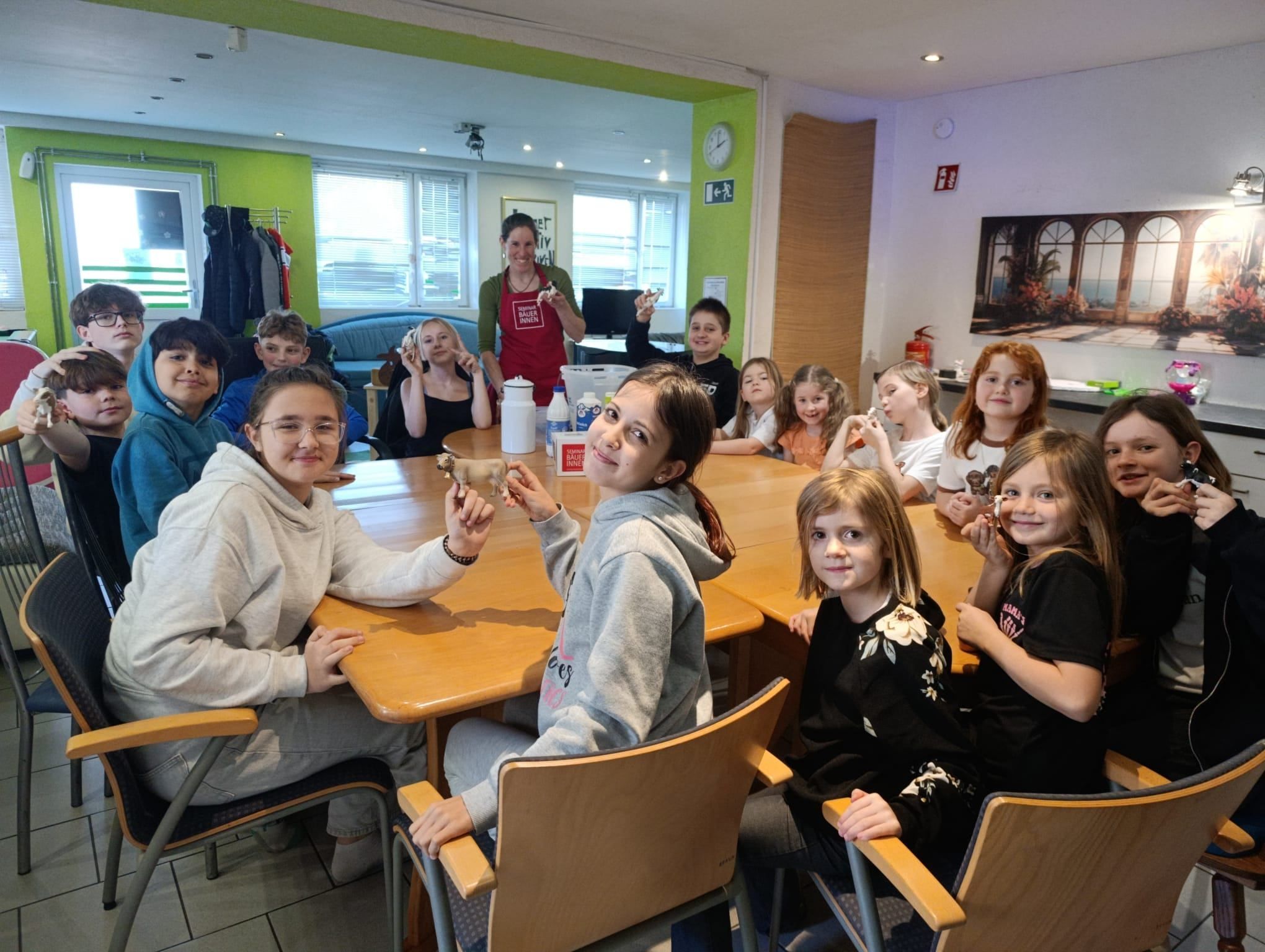 Eine Gruppe von Kindern und einer erwachsenen Frau sitzen um einen Tisch in einem Klassenzimmer, halten kleine Objekte und lächeln. Die Frau trägt eine Schürze und hält einen Behälter. Der Raum hat eine grüne Wand und eine Uhr.