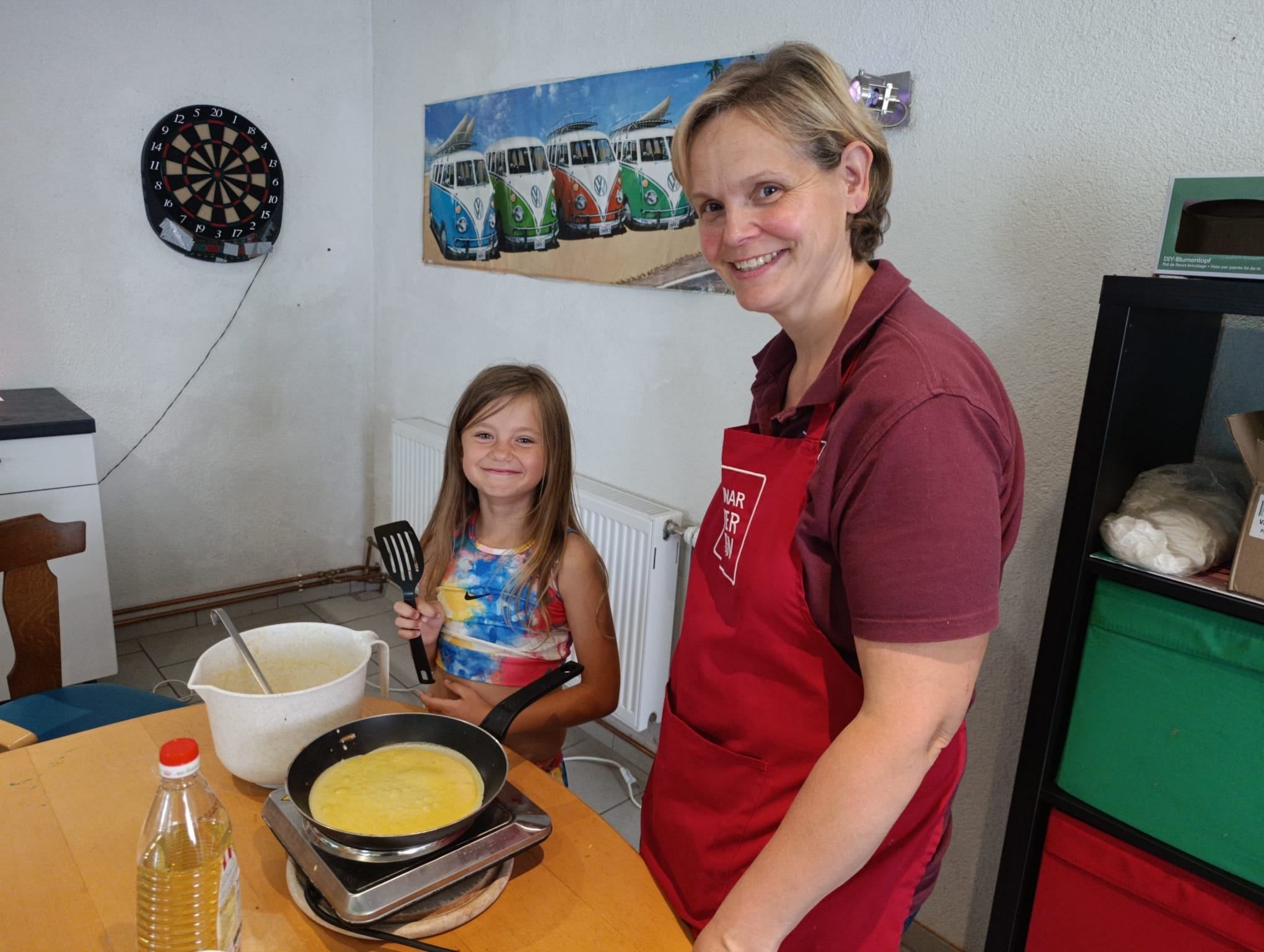 Eine Frau und ein junges Mädchen kochen gemeinsam in einer Küche. Die Frau trägt ein rotes Schürzen und lächelt, während das Mädchen einen Kochlöffel hält. Sie kochen in einer Pfanne auf einem Herd.