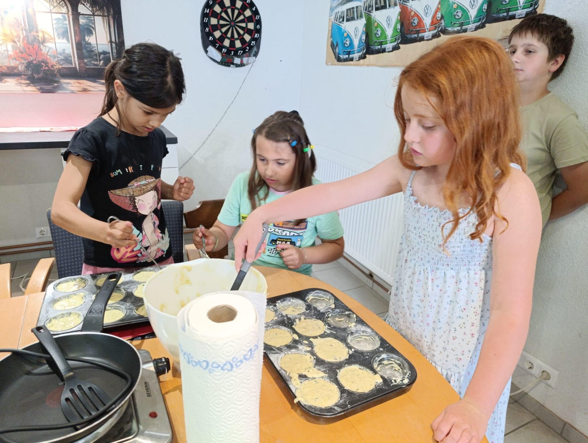 Drei junge Mädchen sind in einer Küche und bereiten sich zum Backen vor. Ein Mädchen rührt Teig in einer Schüssel, während ein anderes den Teig in Muffinformen füllt. Das dritte Mädchen greift nach einer Muffinform. Die Küche hat ein Dartbrett und ein VW-Bulli-Poster an der Wand.