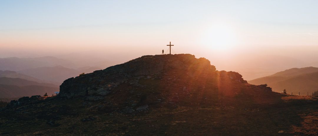 Eine Person steht auf einem felsigen Berggipfel mit einem Kreuz bei Sonnenuntergang. Der Himmel ist klar und orange.