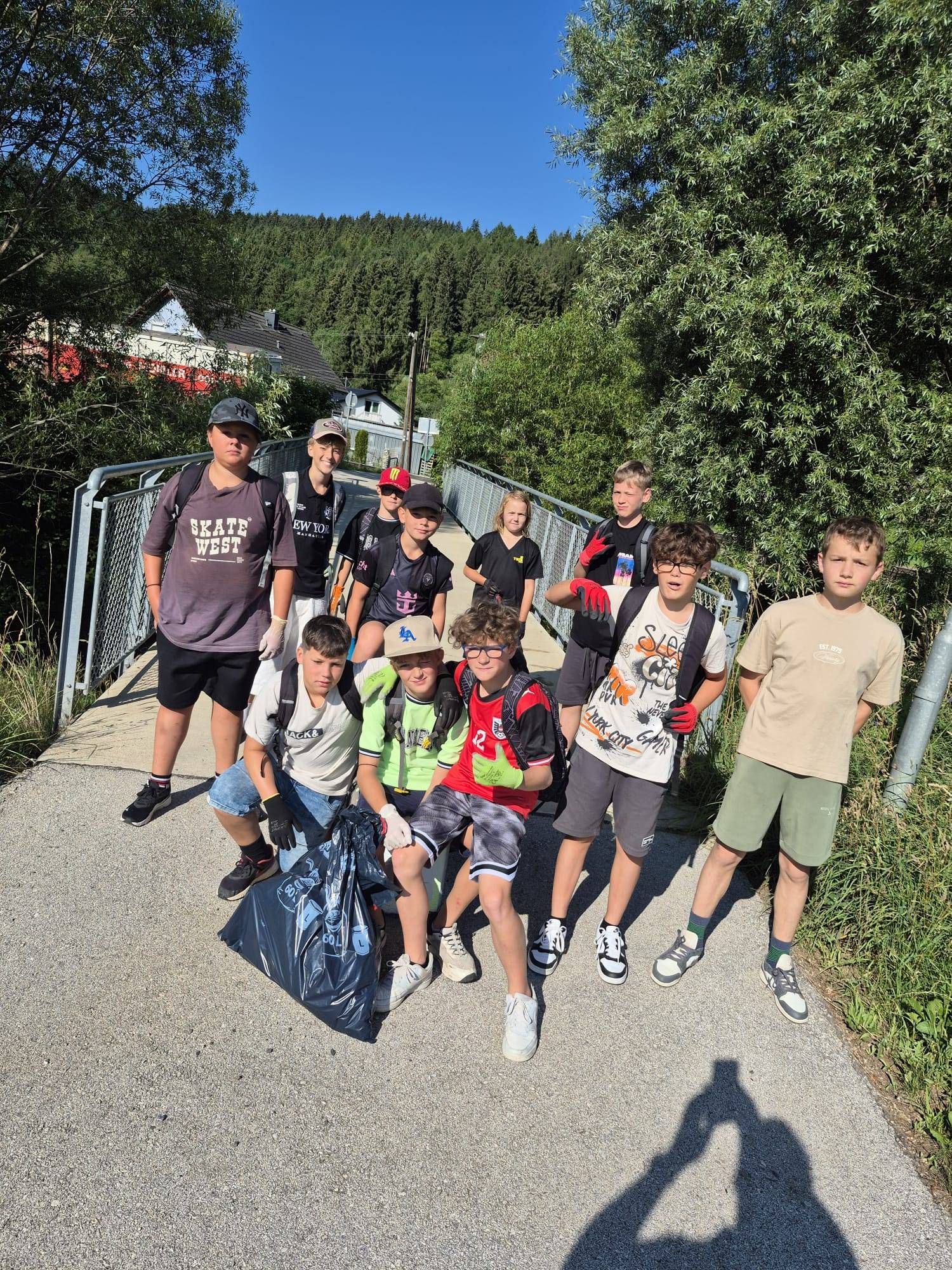 Eine Gruppe von Kindern, die Handschuhe tragen und Müllsäcke mit sich führen, steht auf einem Betonweg. Einige tragen Hüte und Brillen. Hinter ihnen befinden sich Bäume und ein Haus.