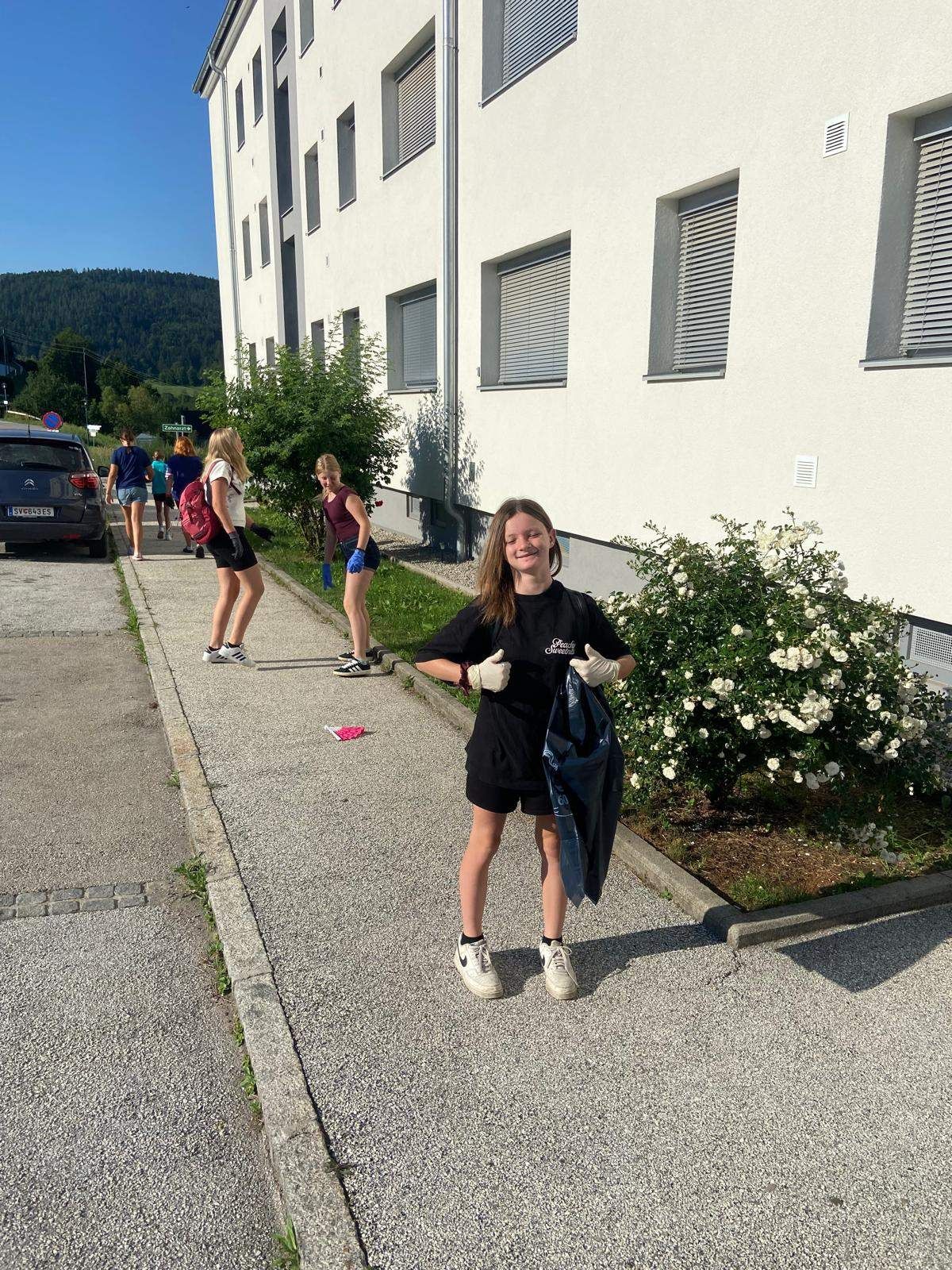 Eine junge Frau steht auf einem Bürgersteig in der Nähe eines Gebäudes, hält eine blaue Tasche in der Hand, während andere vorbeigehen und ein Auto geparkt ist.