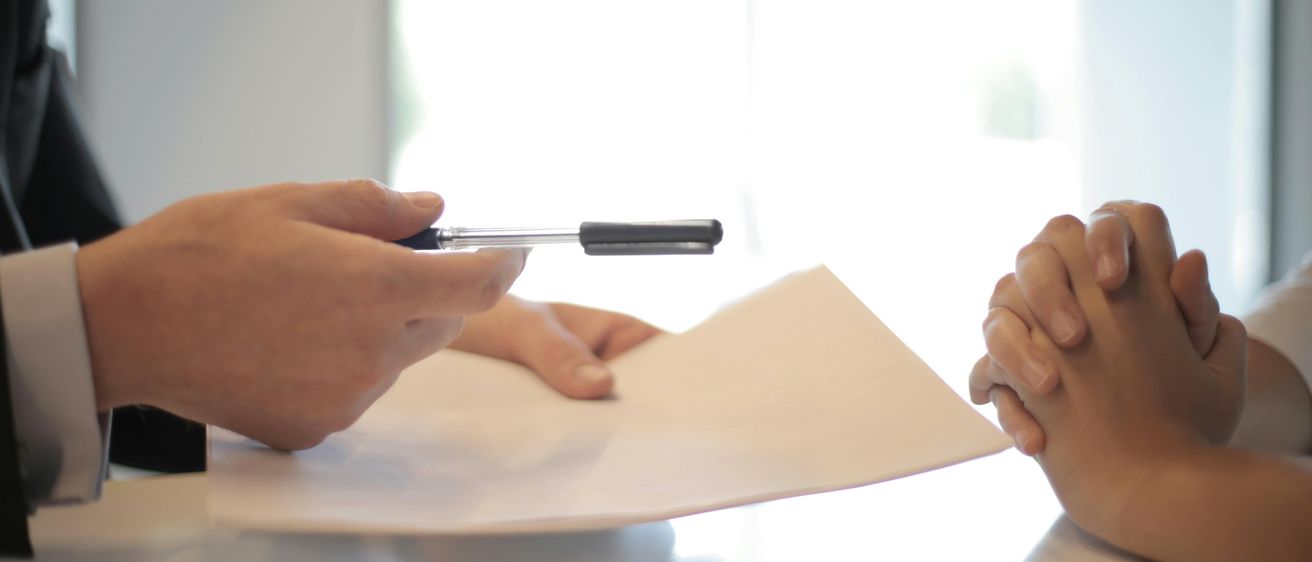 Zwei Hände halten einen Stift und ein Blatt Papier auf einem Tisch, mit einem Fenster und einer Außensicht im Hintergrund.