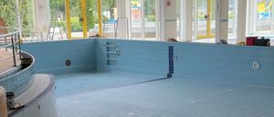 Ein Hallenbad mit blauen Fliesen und gelben Wänden, mit einer Sprungbrett links und einer Leiter rechts. Der Bereich ist leer, mit einigen Gegenständen in der Nähe des Beckens.