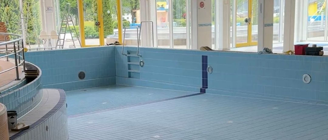 Ein Hallenbad mit blauen Fliesen und gelben Wänden, mit einer Sprungbrett links und einer Leiter rechts. Der Bereich ist leer, mit einigen Gegenständen in der Nähe des Beckens.