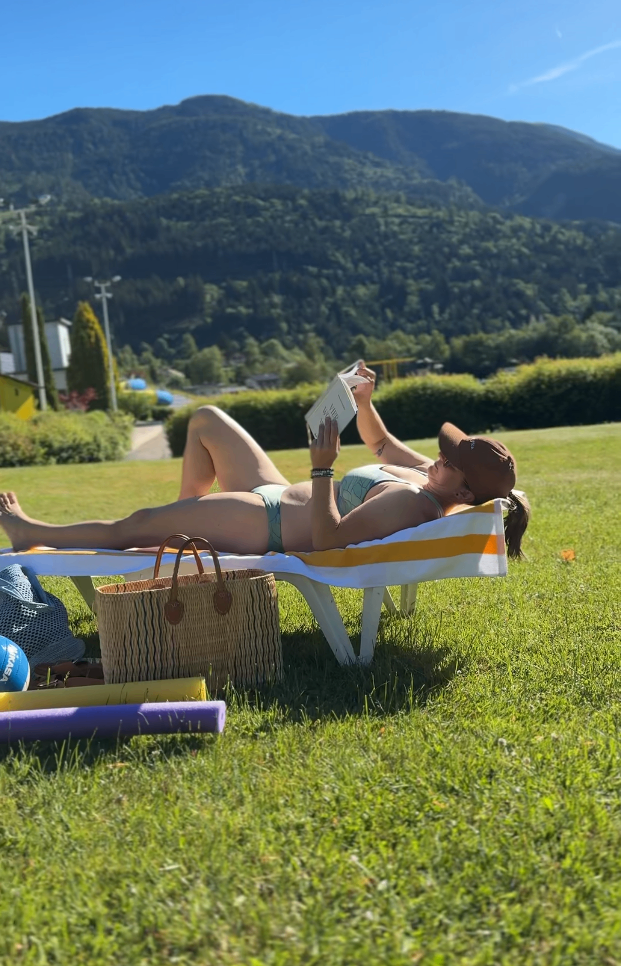 A woman lies on a lounge chair on a grassy field, reading a book. She wears a bikini and a hat. Nearby, there is a basket and a ball. In the background, mountains and a building are visible.