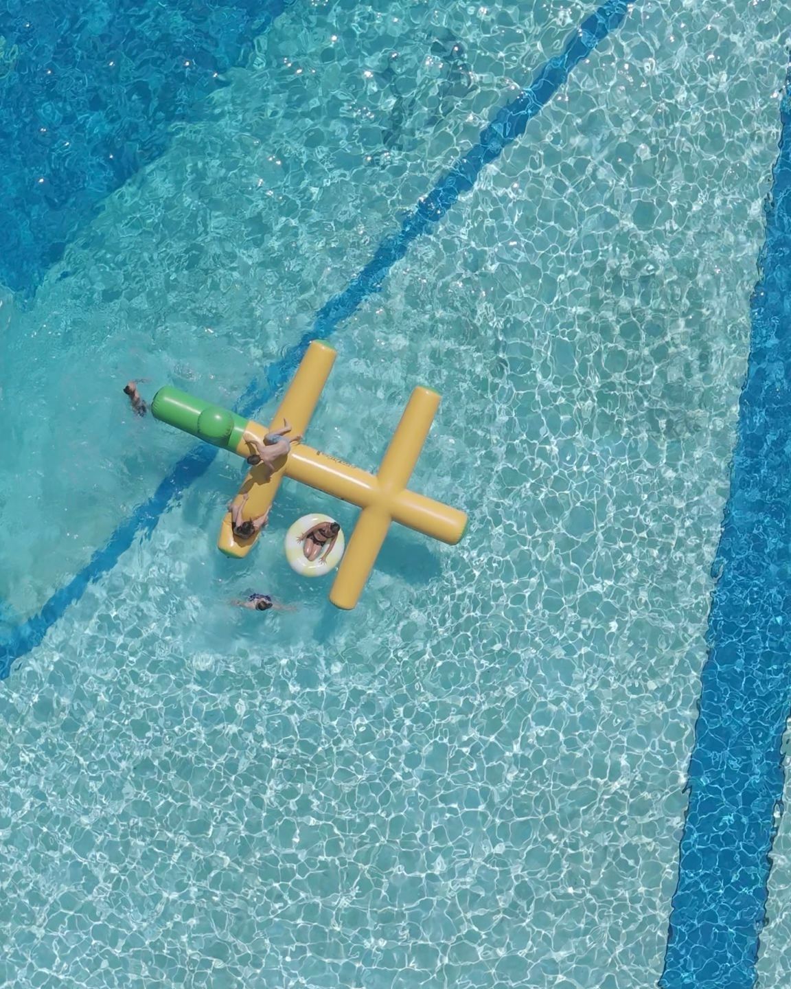 Eine Luftaufnahme zeigt einen Pool mit einem gelben X-förmigen aufblasbaren Schwimmkörper. Mehrere Menschen vergnügen sich darauf und im Wasser.