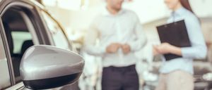 Ein Mann und eine Frau diskutieren über den Verkauf eines Autos in einem Autohaus. Der Mann zeigt auf das Auto, und die Frau hält eine Mappe.