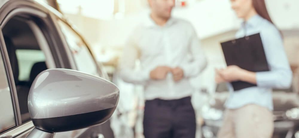 Ein Mann und eine Frau diskutieren über den Verkauf eines Autos in einem Autohaus. Der Mann zeigt auf das Auto, und die Frau hält eine Mappe.