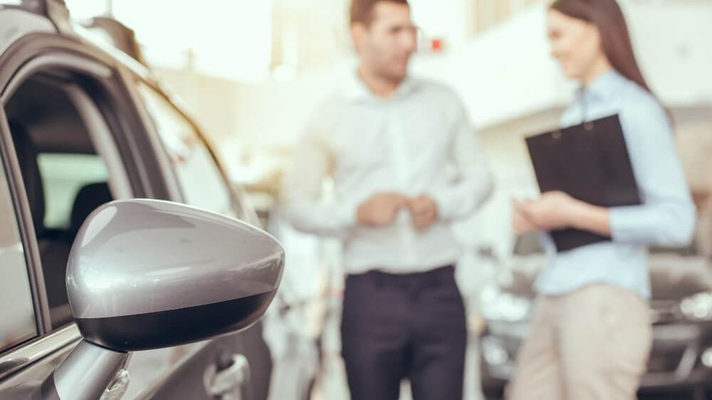 Ein Mann und eine Frau diskutieren über den Verkauf eines Autos in einem Autohaus. Der Mann zeigt auf das Auto, und die Frau hält eine Mappe.