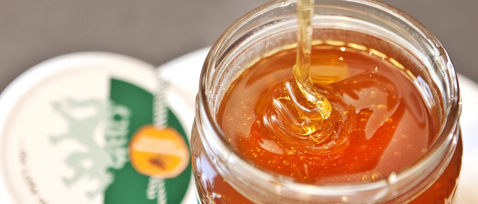 Eine Nahaufnahme eines Glases Honig mit einem dicken, goldenen Strom, der von einem Löffel in das Glas tropft, umgeben von einem unscharfen Hintergrund mit einem grünen und orangefarbenen Logo.