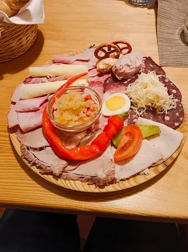 Ein hölzerner Teller hat verschiedene Fleischsorten, Gemüse und Beilagen darauf. Im Hintergrund befindet sich ein Korb mit Brot.