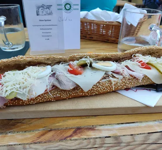 Ein langes Sandwich mit Schinken, Käse, Eiern, Tomaten und Gurken liegt auf einem Holzschneidebrett. Zwei Gläser Wasser und ein Korb stehen auf dem Tisch.