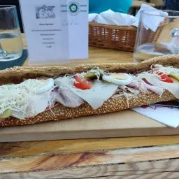 Ein langes Sandwich mit Schinken, Käse, Eiern, Tomaten und Gurken liegt auf einem Holzschneidebrett. Zwei Gläser Wasser und ein Korb stehen auf dem Tisch.