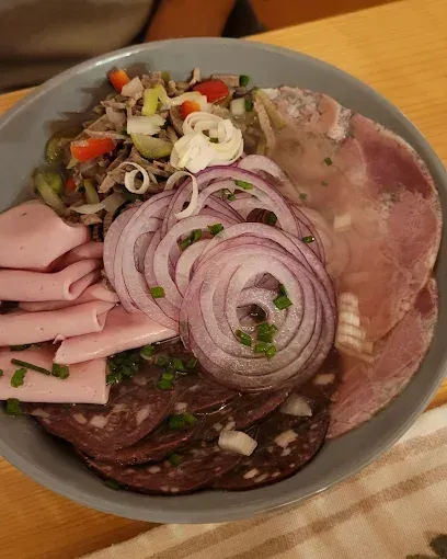 Eine Schüssel auf einem Holztisch gefüllt mit verschiedenen Fleischsorten, darunter geschnittener Schinken und Salami, Zwiebeln und Gemüse.