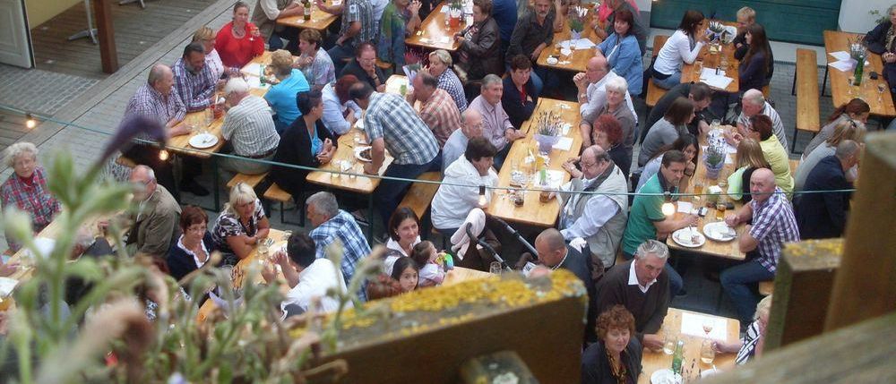 Eine große Gruppe von Menschen sitzt draußen an Tischen, möglicherweise für ein gemeinschaftliches Treffen. Über ihnen hängen leuchtende Lichterketten.