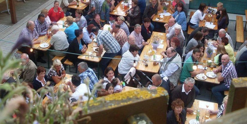 Eine große Gruppe von Menschen sitzt draußen an Tischen, möglicherweise für ein gemeinschaftliches Treffen. Über ihnen hängen leuchtende Lichterketten.