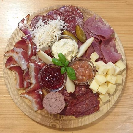 Ein Holzteller mit verschiedenen Fleischsorten, Käse und Beilagen. Die Fleischsorten umfassen Salami, Speck und Wurst. Die Käsesorten sind in Würfeln angeordnet. Zu den Beilagen gehören eine cremige Sauce und eine rote Gelee.
