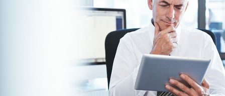 Ein Mann in einem weißen Hemd und einer Krawatte sitzt im Büro und benutzt ein Tablet. Er sieht konzentriert aus und berührt sein Kinn mit der rechten Hand. Hinter ihm sind ein Computermonitor und eine Tastatur zu sehen.