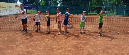 Eine Gruppe von Kindern und einem Trainer stehen auf einem Tennisplatz mit Schlägern, halten gelbe Bälle und tragen Mützen und Turnschuhe. Dahinter ist ein Tennisnetz und Bäume zu sehen.