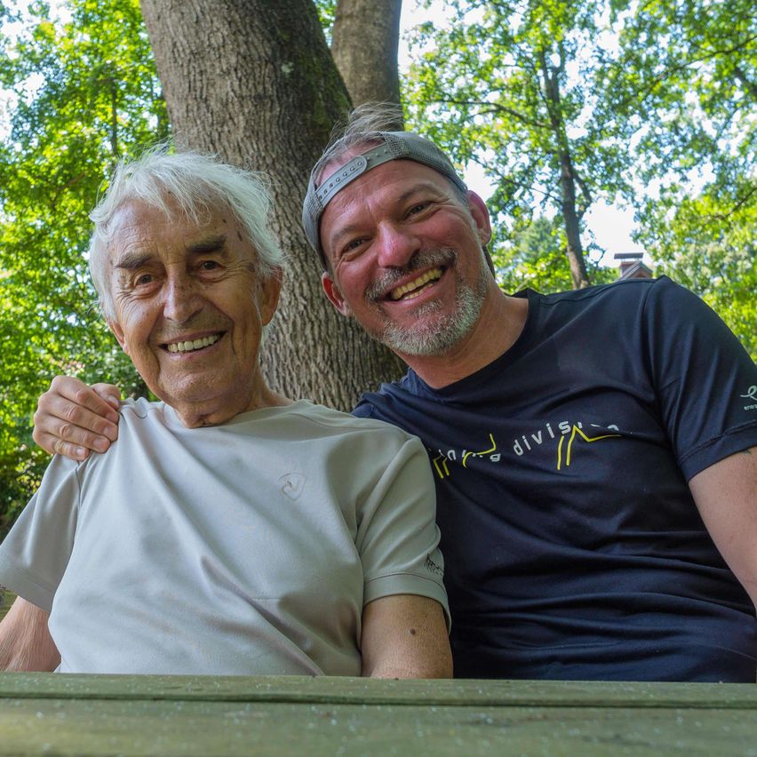 Zwei Männer lächeln und posieren für ein Foto in einem Park. Der ältere Mann hat seinen Arm um den jüngeren Mann gelegt. Sie tragen beide Freizeitkleidung.