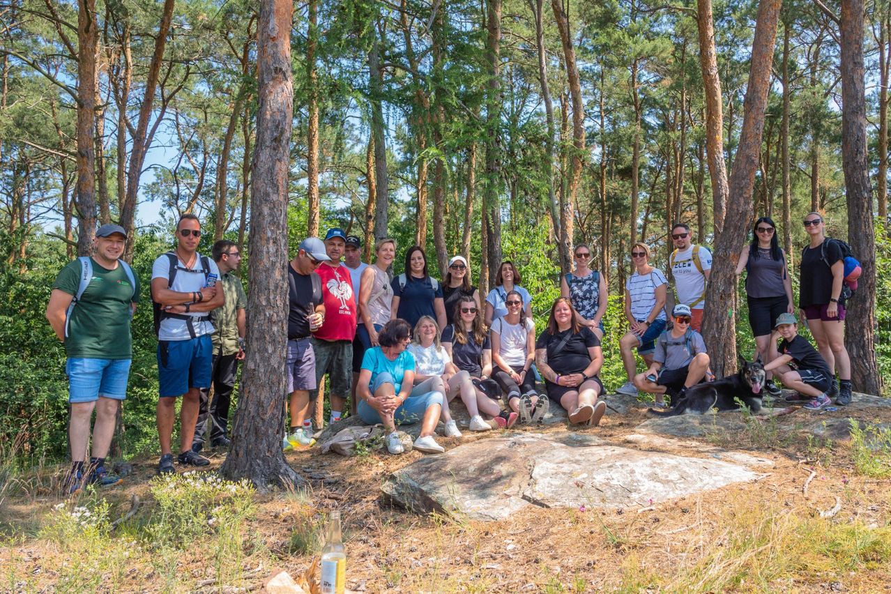 Eine Gruppe von Menschen, einige stehend und einige sitzend, posiert für ein Foto im Wald. Bäume umgeben sie, und einige tragen Sonnenbrillen. Ein Hund liegt auf dem Boden.