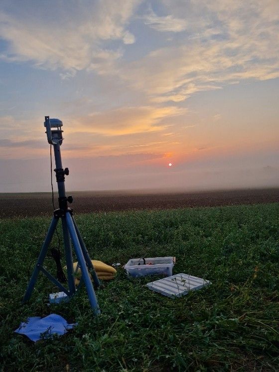 Ein Stativ mit einem Vermessungsinstrument steht in einem Grasfeld. Die Sonne geht in der Ferne unter und taucht alles in ein warmes Licht. Verschiedene Gegenstände sind verstreut.
