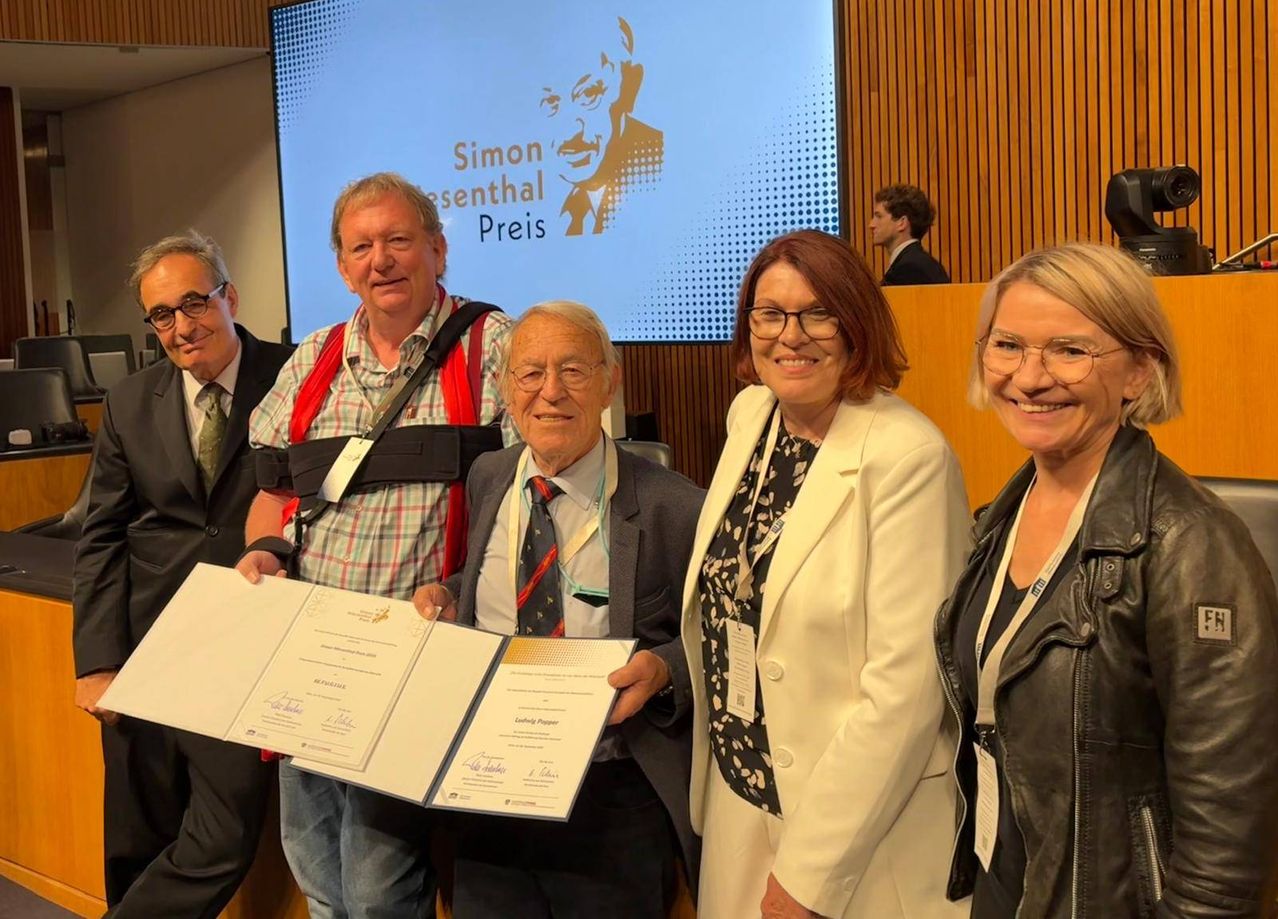 Fünf Personen stehen in einem Raum, lächeln und posieren für ein Foto. Ein Mann hält ein Zertifikat. Dahinter zeigt eine Projektionsleinwand ein Bild eines Mannes und die Worte 'Simon-Esenthel Preis'.