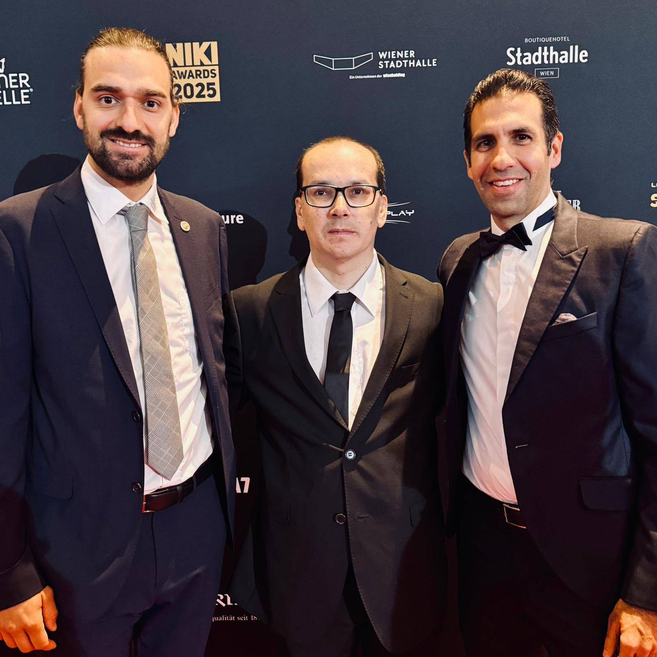 Drei Männer stehen zusammen auf einem roten Teppich. Sie sind in formeller Kleidung gekleidet, einer trägt eine Brille. Der Hintergrund zeigt Logos und Text auf Deutsch.