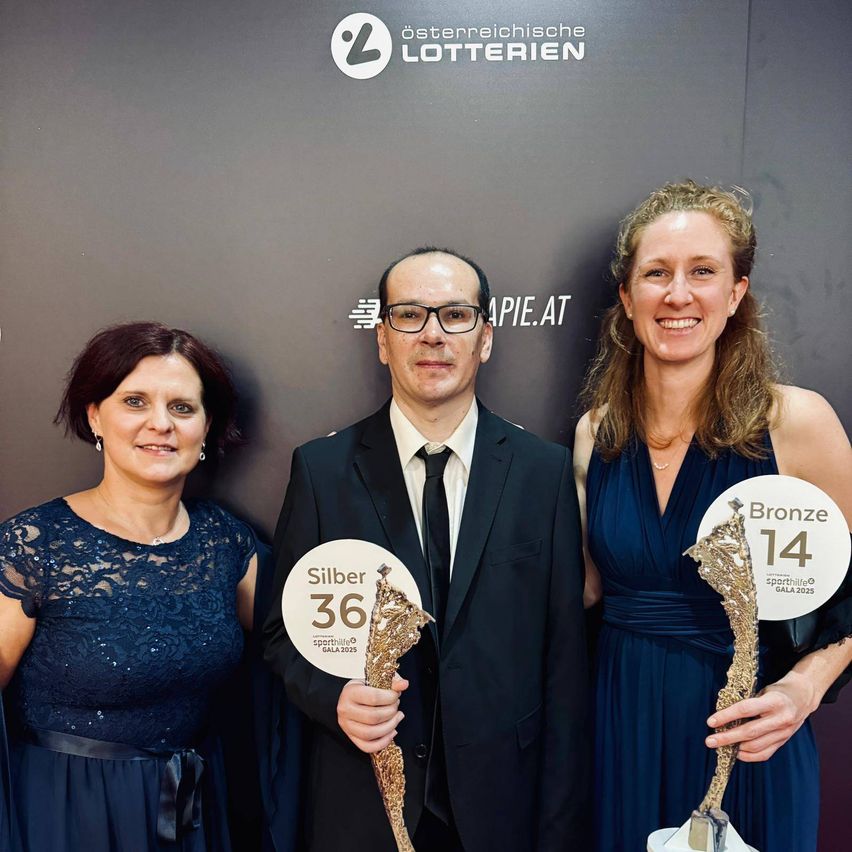 Drei Menschen, zwei Frauen und ein Mann, stehen zusammen und halten Trophäen. Der Mann hält eine Silbertrophäe, während die beiden Frauen Bronzetrophäen halten. Hinter ihnen befindet sich eine Wand mit Logos und Text.